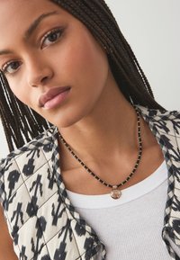Black and gold beaded necklace with a round pendant. Model wears a patterned vest over a white ribbed tank top. Close-up on the neckline.