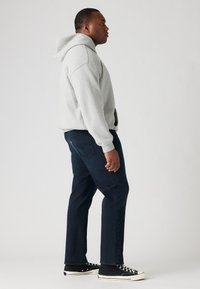 Grijze fleece hoodie met een voorzak, gecombineerd met donkerblauwe spijkerbroek. Zwarte hoge sneakers met witte zolen zichtbaar aan de onderkant.