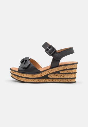 Sorte kilehæl sandaler med en bueaccent, justerbar spænde rem, korkstruktureret sål og tekstureret detaljering på kiledesignet.