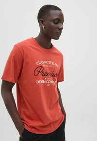 Raudona medvilnė mišrainė marškinėliai su apvali iškirpte, trumpomis rankovėmis ir juodu spaudiniu, kuriame yra "Premium Denim Company" ir įsteigimo detalės.
