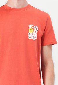 Bomuld t-shirt i koral, med hvid tekst "F*ck Off Mondays" og et gult smiley-ansigt på venstre brystområde. Korte ærmer.