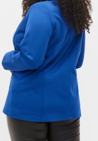 Blå figursydd blazer i texturerad tyg, med en skräddarsydd design och subtila sömdetaljer. Matchad med glänsande svarta byxor.