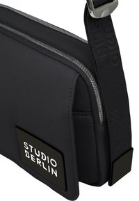 Borsa a spalla in nylon nero con doppia cerniera, una tracolla texturizzata e una toppa rettangolare con "STUDIO BERLIN" in bold lettere bianche.