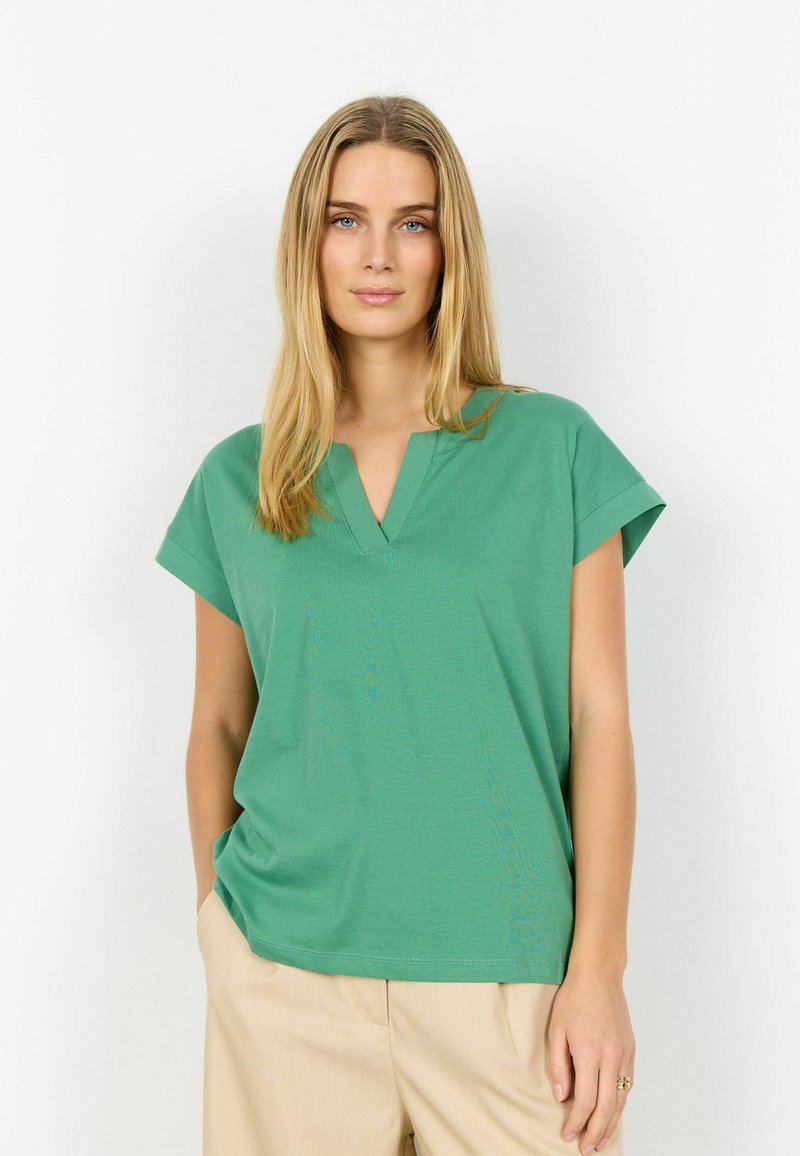 Femme aux longs cheveux blonds, portant un haut vert à manches courtes avec col en V et un pantalon beige, debout avec une main dans la poche.