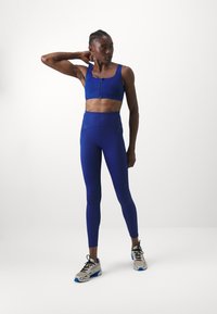 Sinine spordibrasi ees oleva tõmblukuga, millele on paariud kõrge vöökohaga sinised legginsid. Mudel kannab halli spordijalatsit siniste aktsentidega. Sile tekstuur.