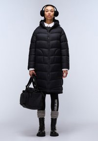 Lange zwarte pufferjas met een capuchon, ritssluiting en gequilte design. Zwarte legging met een logo en grijze gebreide beenwarmers. Zwarte laarzen en tas.