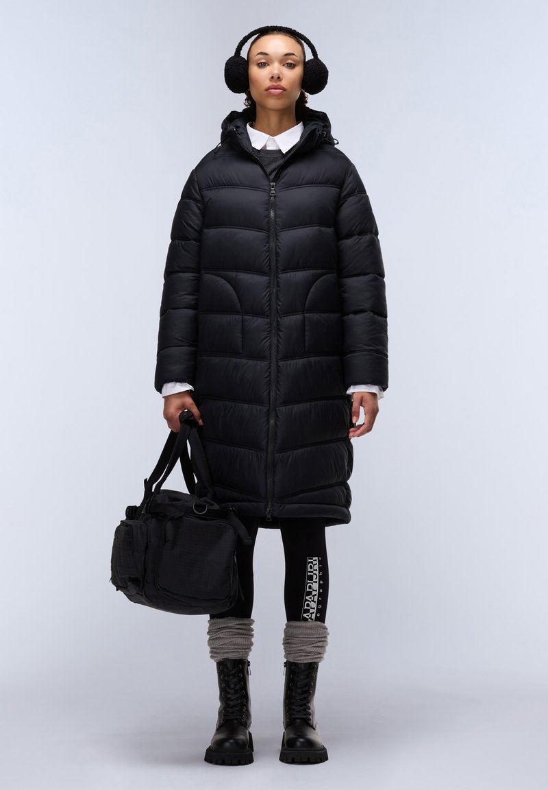 Lange zwarte pufferjas met een capuchon, ritssluiting en gequilte design. Zwarte legging met een logo en grijze gebreide beenwarmers. Zwarte laarzen en tas.