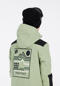 Grüne wasserdichte Jacke mit schwarzen Akzenten, die ein geometrisches Bergdesign und Logo auf dem Rücken aufweist. Weiche, schwarze Strickmütze getragen.