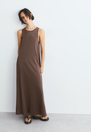 Femme aux cheveux courts et foncés, debout appuyée contre un mur, portant une robe longue marron sans manches et des sandales à plateforme noires.