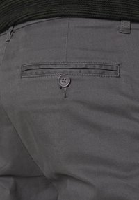 Los pantalones grises presentan un bolsillo trasero con apertura horizontal y un cierre de botón negro. La tela tiene una textura suave y un brillo sutil.