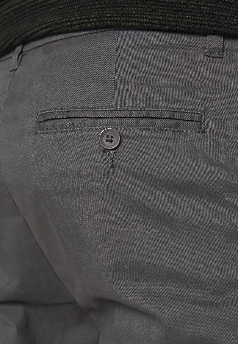 Los pantalones grises presentan un bolsillo trasero con apertura horizontal y un cierre de botón negro. La tela tiene una textura suave y un brillo sutil.