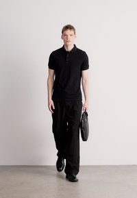 Melnā polo krekls ar apkakli, kopā ar vaļīgām melnām biksēm. Tur melnu ādas somu, valkā melnus apavus. Minimālisma dizains, neitrālas krāsas.