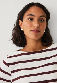 Witte longsleeve top met bordeauxrode horizontale strepen, ronde halslijn en een relaxte pasvorm. Model draagt gouden cilinder oorbellen.