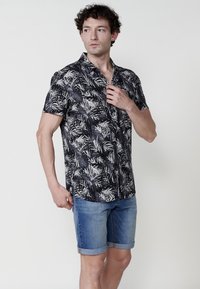Camisa negra de manga corta con un patrón de hojas tropicales blancas, combinada con pantalones cortos de mezclilla azules. La tela parece ligera y casual.