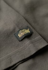 Gråt stof med en glat tekstur har en sort label med gul broderi, der detaljerer et design af en struktur. Synlige stikkanter.