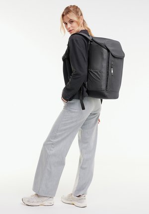 Junge Frau trägt eine schwarze Jacke und hellgraue Hose, trägt einen großen schwarzen Rucksack und steht vor einem schlichten weißen Hintergrund.