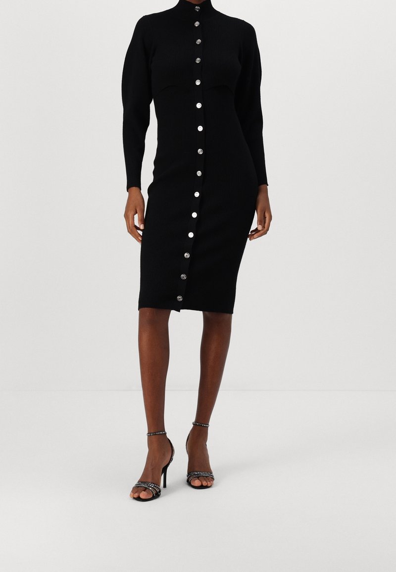 Femme portant une robe midi noire ajustée à manches longues avec des boutons argentés sur le devant, associée à des sandales noires à talons hauts et bride à la cheville.