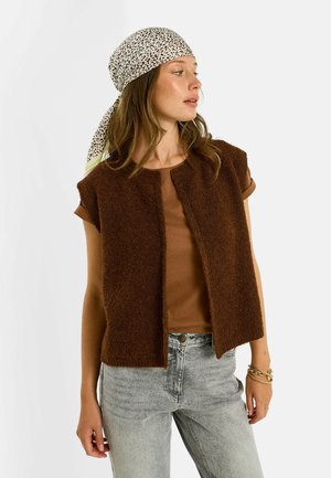 Mujer con cabello castaño claro lleva un pañuelo blanco con estampado de leopardo, chaleco sin mangas marrón y peludo, camisa marrón, vaqueros gris claro y pulseras doradas.