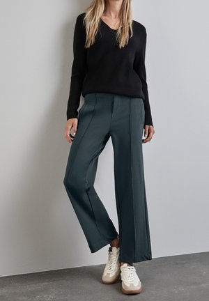 Vrouw die een zwarte top met lange mouwen, donkergroene wijde broek en witte sneakers draagt, staand tegen een lichtgrijze muur.