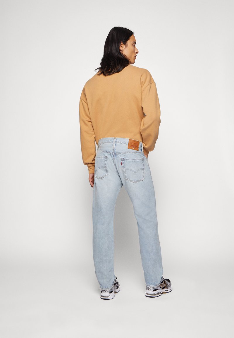 Helle blaue Denim-Jeans mit lockerer Passform und Lederpatch. Kombiniert mit einem hellbraunen Sweatshirt. Sneakers sind am unteren Ende sichtbar.