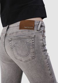 Jean en denim gris à coupe slim, comportant une patch en cuir brun avec l'inscription "True Religion" et deux poches arrière avec des accents de couture.