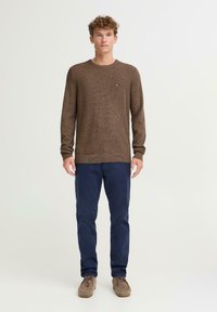 Brązowy sweter o fakturze z okrągłym dekoltem i dopasowanymi mankietami, zestawiony z granatowymi spodniami i jasnobrązowymi mokasynami. Sweter ma małe logo.