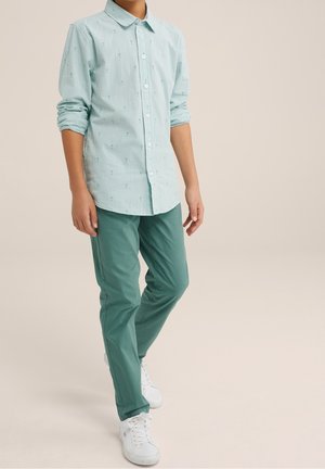 Chemise à manches longues rayée bleu clair avec un col boutonné, associée à un pantalon vert et des baskets blanches, debout devant un fond neutre.