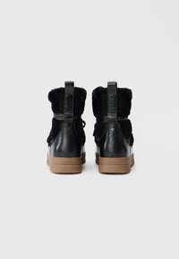 Une paire de bottes noires en cuir et en fourrure, avec des tirants de traction, des lacets et des semelles en caoutchouc marron, montrées de dos sur un fond blanc uni.