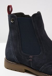 Bottines en daim bleu marine foncé avec des panneaux élastiques sur les côtés et un tirant bordeaux. Elles présentent une surface texturée et une semelle en caoutchouc brun.