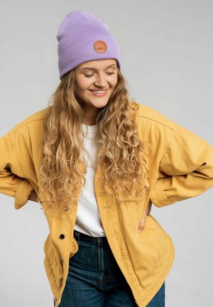 Żółta, oversize'owa kurtka na białej koszulce, w zestawie z ciemnoniebieskimi dżinsami. Jasnofioletowa czapka beanie z okrągłą skórzaną łatką z przodu.