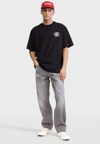 Schwarzes Baumwoll-T-Shirt mit grafischem Druck auf der linken Brust, kombiniert mit hellgrauen Jeans und weißen Sneakers sowie einer roten Kappe.
