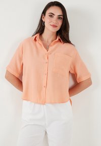 Short-sleeve, button-up shirt in light peach linen fabric with a single chest pocket and a cropped hemline, paired with white pants.