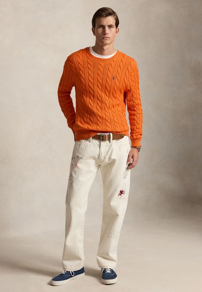 Ung mann som står iført en oransje kabelstrikket genser, hvite mønstrete bukser, brun belte, blå joggesko og klokke på venstre håndledd.