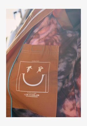 Binnenkant van een oranje jack met een mesh-textuur. Bevat een witte graphic van een glimlachend gezicht met twee figuren, gelabeld "LOS ANGELES." Inclusief een ritszak.