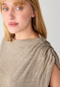 Haut beige en maille à coupe décontractée, avec des épaules froncées et un large décolleté. Le tissu présente une apparence douce et texturée.