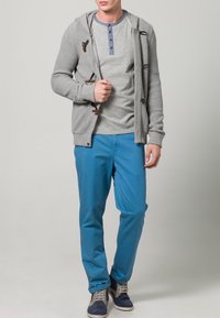 Šedý žebrovaný kardigan s kapucí a dřevěnými knoflíky, nošený přes šedé tričko henley, doplněný modrými kalhotami a tmavě modrými botami.