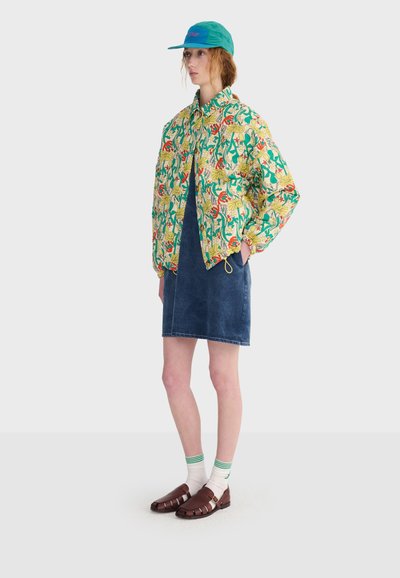 Femme portant une casquette turquoise, une veste jaune à motif floral, une jupe en jean bleue, des chaussettes blanches à rayures vertes et des sandales tressées marron, debout de profil.