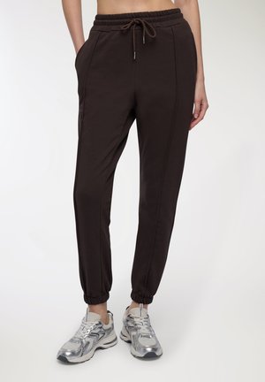 Persona che indossa pantaloni jogger marrone scuro con vita e polsini elasticizzati, abbinati a sneaker atletiche argento e bianche su sfondo bianco.