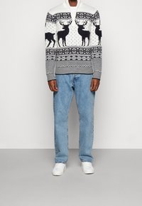 Stickad tröja med dragkedja i nacken, vit basfärg, svarta renar och snöflingemönster, tillsammans med blå jeans och vita sneakers.