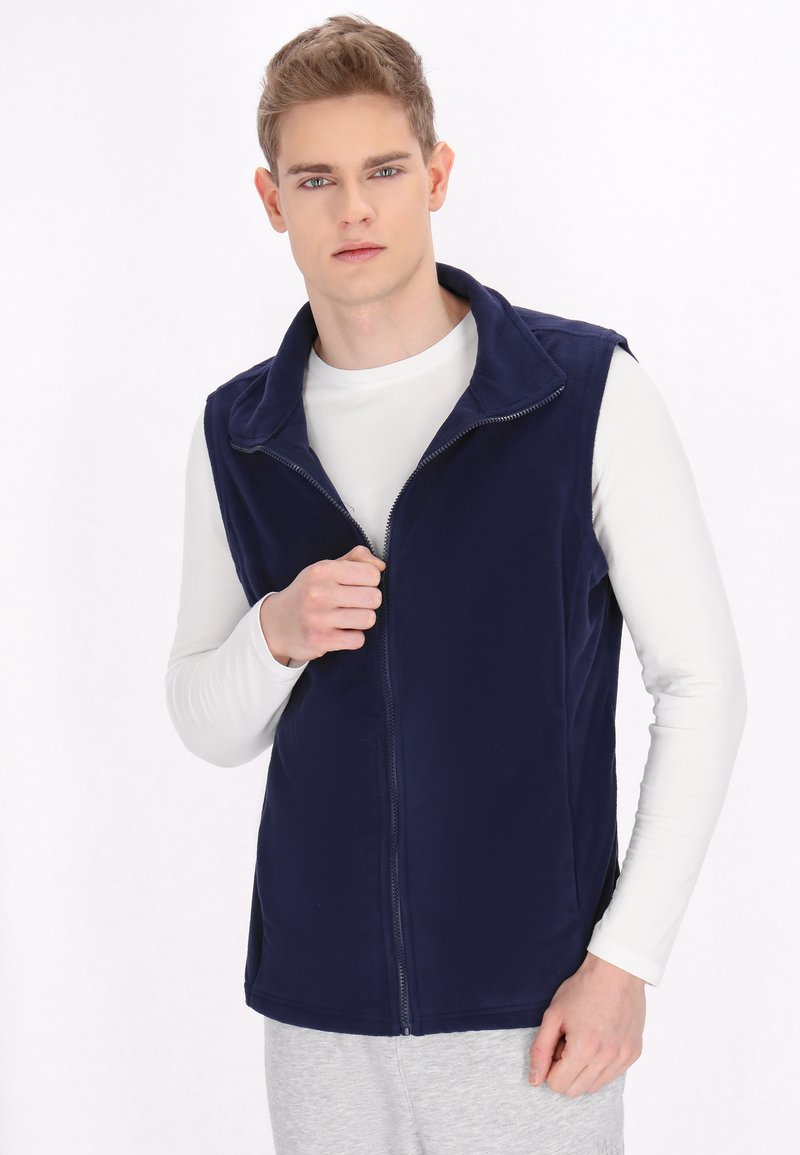 Námornícky modrá fleecová vesta so zipsom vpredu, preloženým golierom a bezrukávovým dizajnom, nosená cez bielu košeľu s dlhým rukávom.