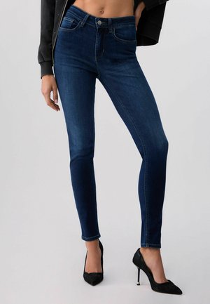 Frau trägt dunkelblaue Skinny-Jeans, schwarze High Heels und eine kurze schwarze Jacke, steht vor einem schlichten hellen Hintergrund.