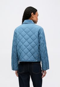 Kvinde iført en blå quiltet jakke og mørke jeans, vist fra ryggen mod en ensfarvet hvid baggrund.