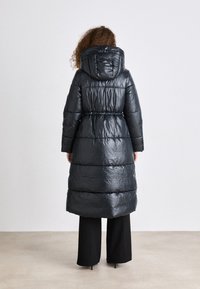 Casaco puffer preto com capuz, apresentando uma cintura ajustada, textura acolchoada e um design longo, usado sobre calças pretas.