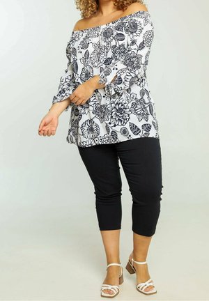 Schulterfreies Oberteil mit schwarzem Blumenmuster auf weißem Hintergrund; kombiniert mit schwarzen, knöchellangen Hosen und weißen, schmalen Sandalen mit Absatz.