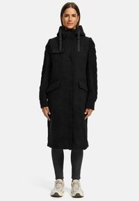 Black long coat with a hood, featuring woven textured sleeves, front pockets, and a zip-up closure. Smooth fabric with contrasting knit accents.