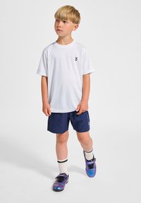 Vit idrottströja med svart logotyp, tillsammans med marinblå shorts. Bär på lila skor och vita strumpor med svarta ränder. Barn står.