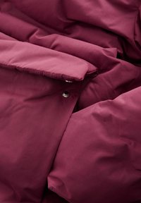 Burgundska posteljnina z mehkim, teksturiranim materialom, ki ima gumbasto zapiranje in plastne gube za udobje.