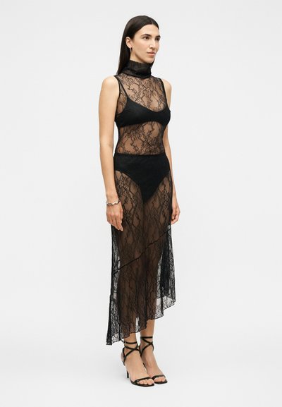Femme portant une robe noire en dentelle sans manches à col montant, des sous-vêtements noirs, et des sandales à talons hauts noires à brides, debout de profil.