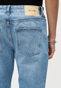 Μεταφορά τζιν από ανοιχτό μπλε ντένιμ, πίσω όψη, με μια ταμπέλα της μάρκας "ONLY & SONS" σε ταμπά χρώμα στη ζώνη και ένα ασημένιο βραχιόλι αλυσίδας στον δεξιό καρπό.