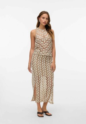 Una donna che indossa un top trasparente senza maniche beige e bianco con motivo zigzag e una gonna midi abbinata con spacco laterale, abbinata a sandali neri aperti sulla punta.
